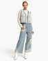 A model stands against a light gray studio background wearing a coordinated outfit consisting of a light blue cropped vest with cream floral embroidery, a cream long-sleeve top, matching wide-leg cropped pants with cream floral trim at the hem and pocket, and white pointed boots.