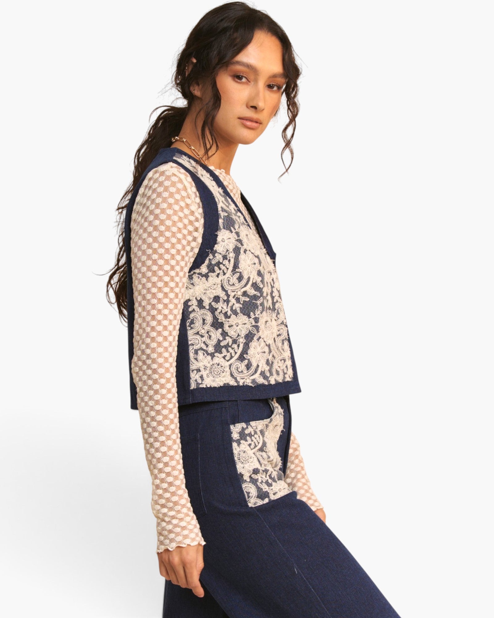 A model wearing a navy blue knit vest with beige and navy damask patterned panels, layered over a beige and navy checkered long-sleeve top and paired with navy blue trousers, against a plain white studio background.