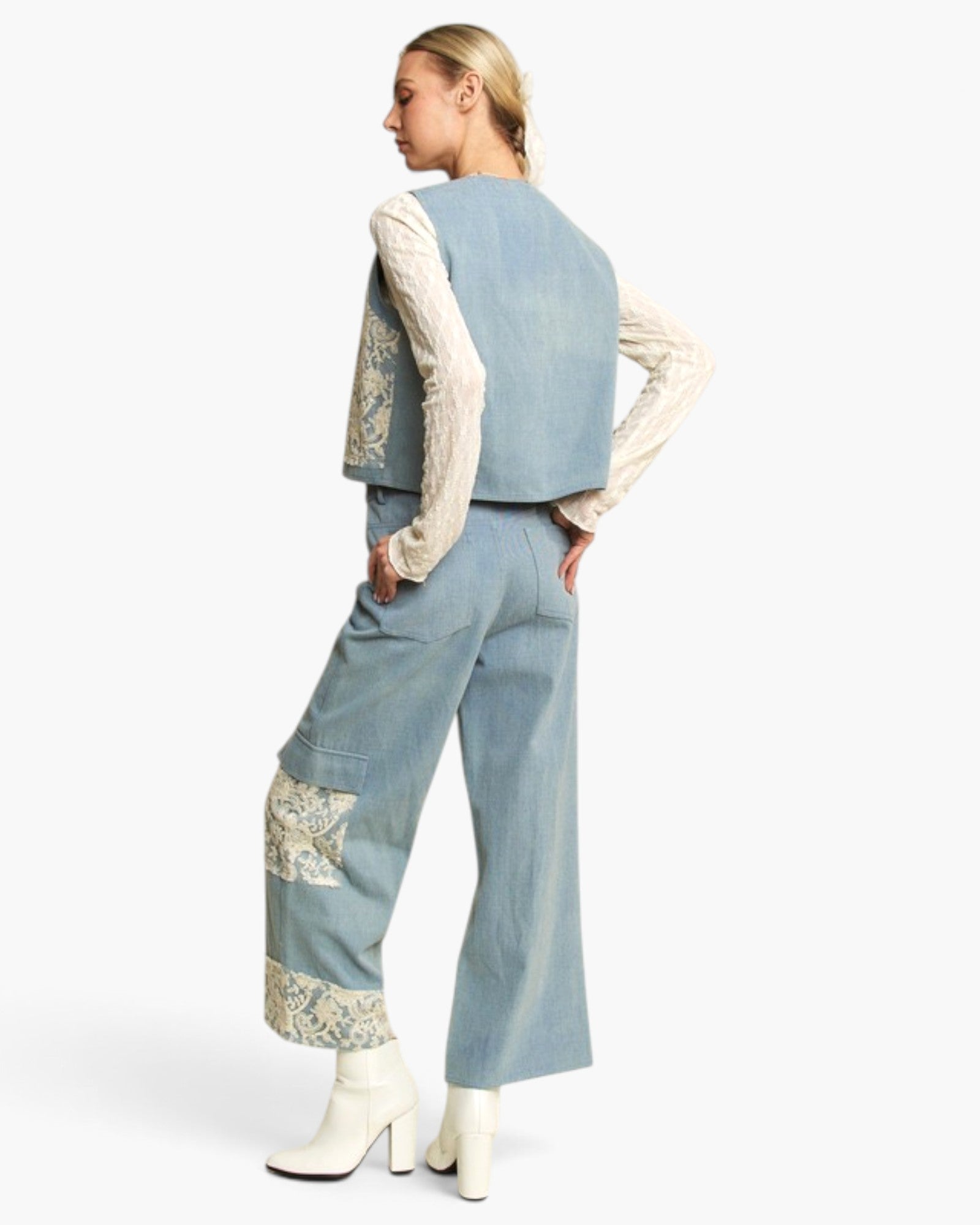 A model shown from the back wearing a light blue denim vest and matching wide-leg jeans with cream lace applique details, layered over a cream lace long-sleeve top and finished with white heeled boots.