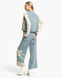 A model shown from the back wearing a light blue denim vest and matching wide-leg jeans with cream lace applique details, layered over a cream lace long-sleeve top and finished with white heeled boots.