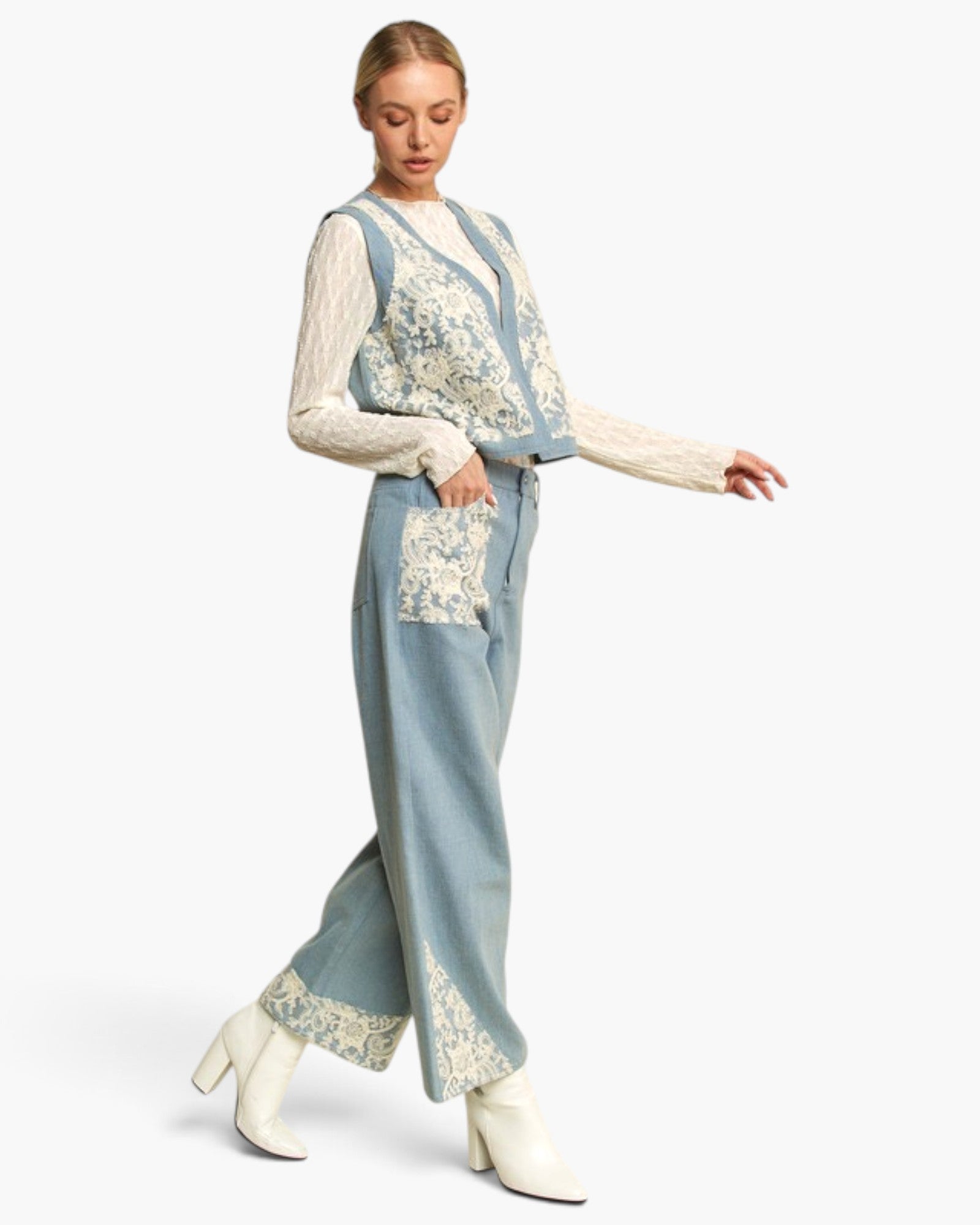 A woman models a light blue denim vest and matching wide-leg trousers set, both decorated with intricate cream floral lace embroidery. She wears a matching cream lace long-sleeve top and white heeled boots against a plain white studio background.
