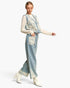 A woman models a light blue denim vest and matching wide-leg trousers set, both decorated with intricate cream floral lace embroidery. She wears a matching cream lace long-sleeve top and white heeled boots against a plain white studio background.