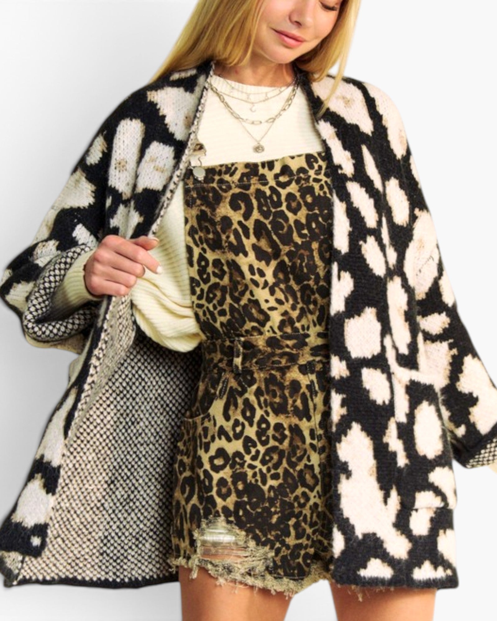 A woman wearing a black and cream ikat pattern knit cardigan over a beige and black leopard print dress, styled with layered gold necklaces against a plain white background.