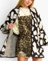 A woman wearing a black and cream ikat pattern knit cardigan over a beige and black leopard print dress, styled with layered gold necklaces against a plain white background.