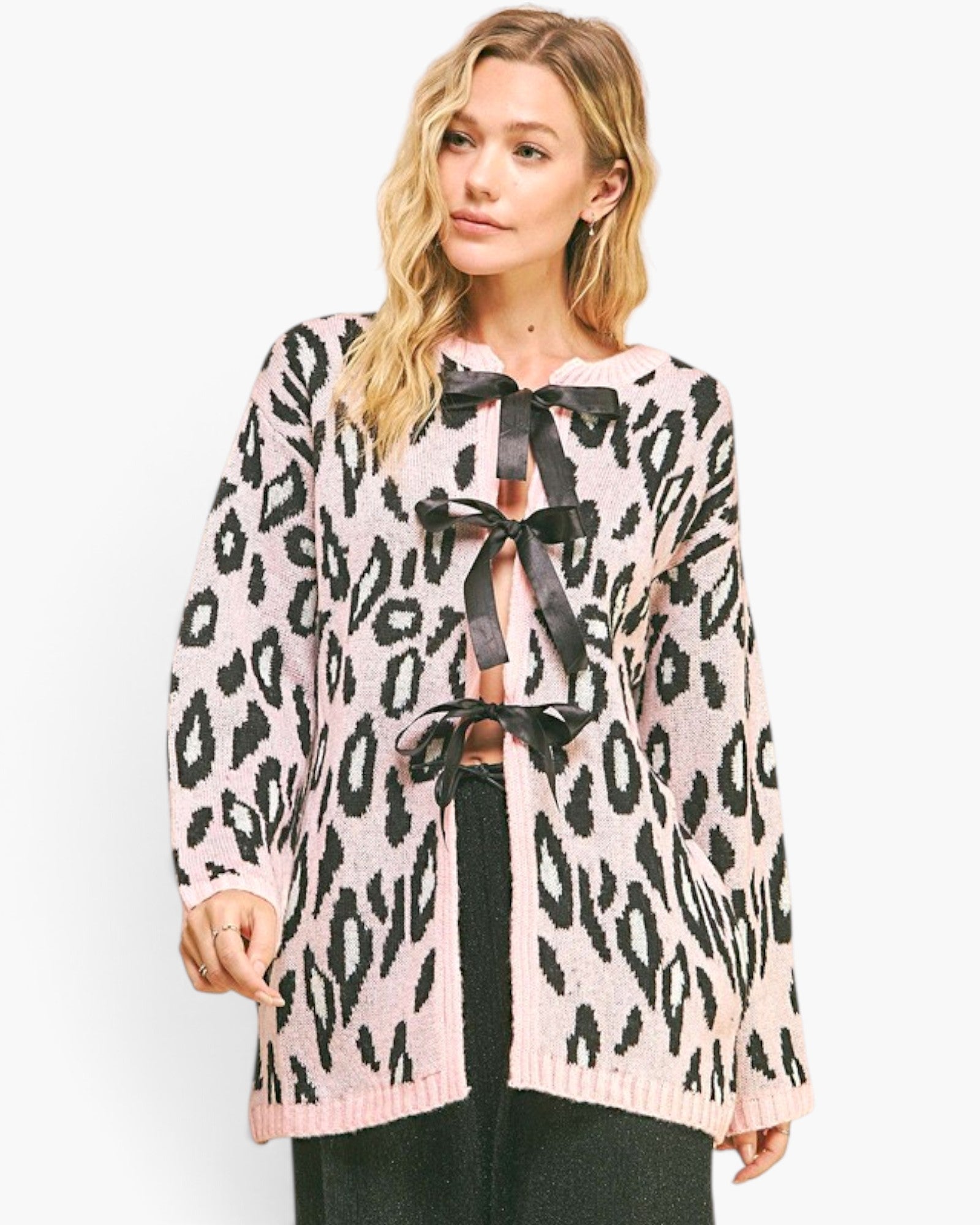 A woman modeling a pink and black leopard print knit cardigan with large black ribbon bows at the front, paired with black pants against a plain white studio background.