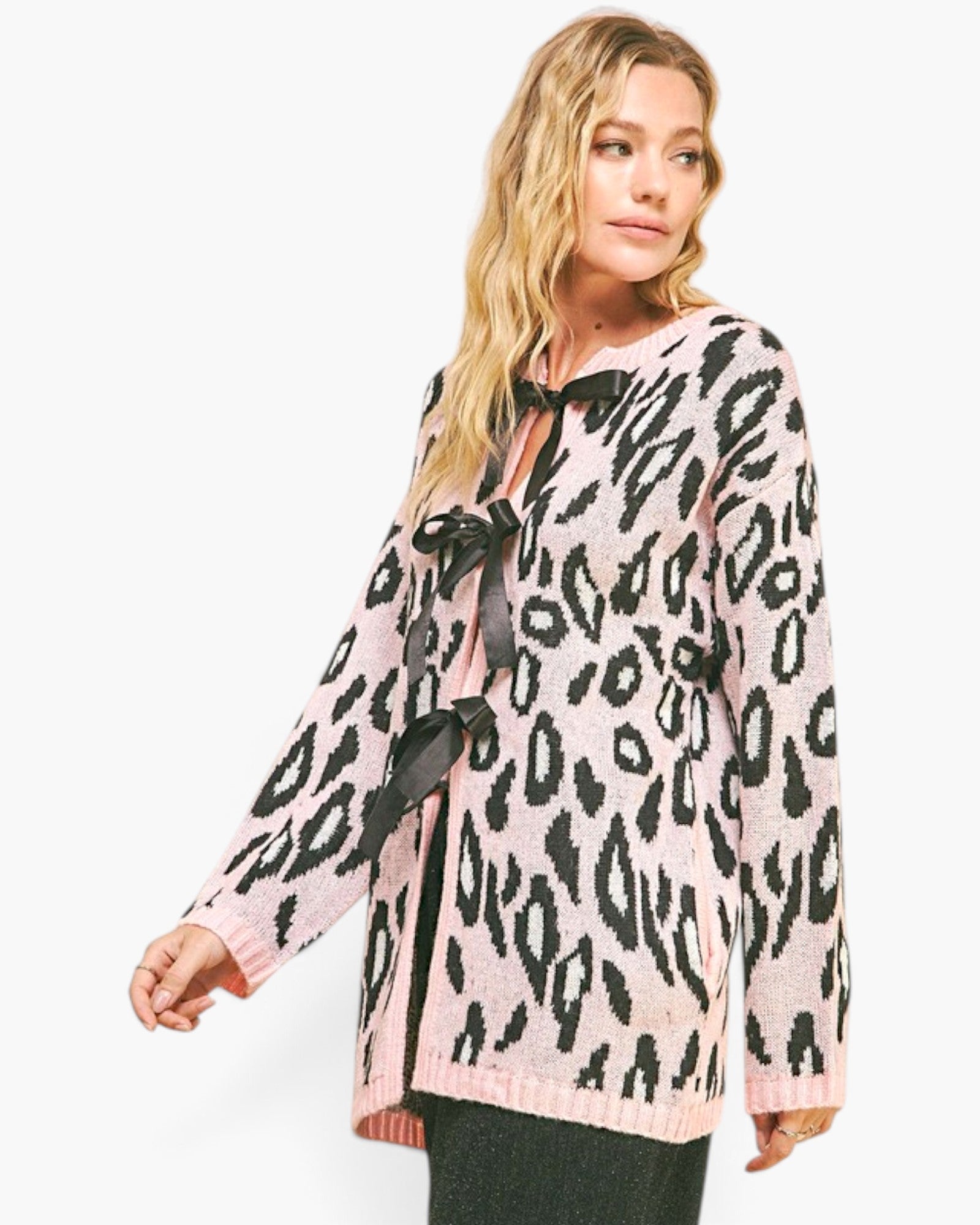 A woman modeling a pink knit cardigan with a black leopard print pattern and black ribbon tie details at the neckline and front, photographed against a plain white studio background.