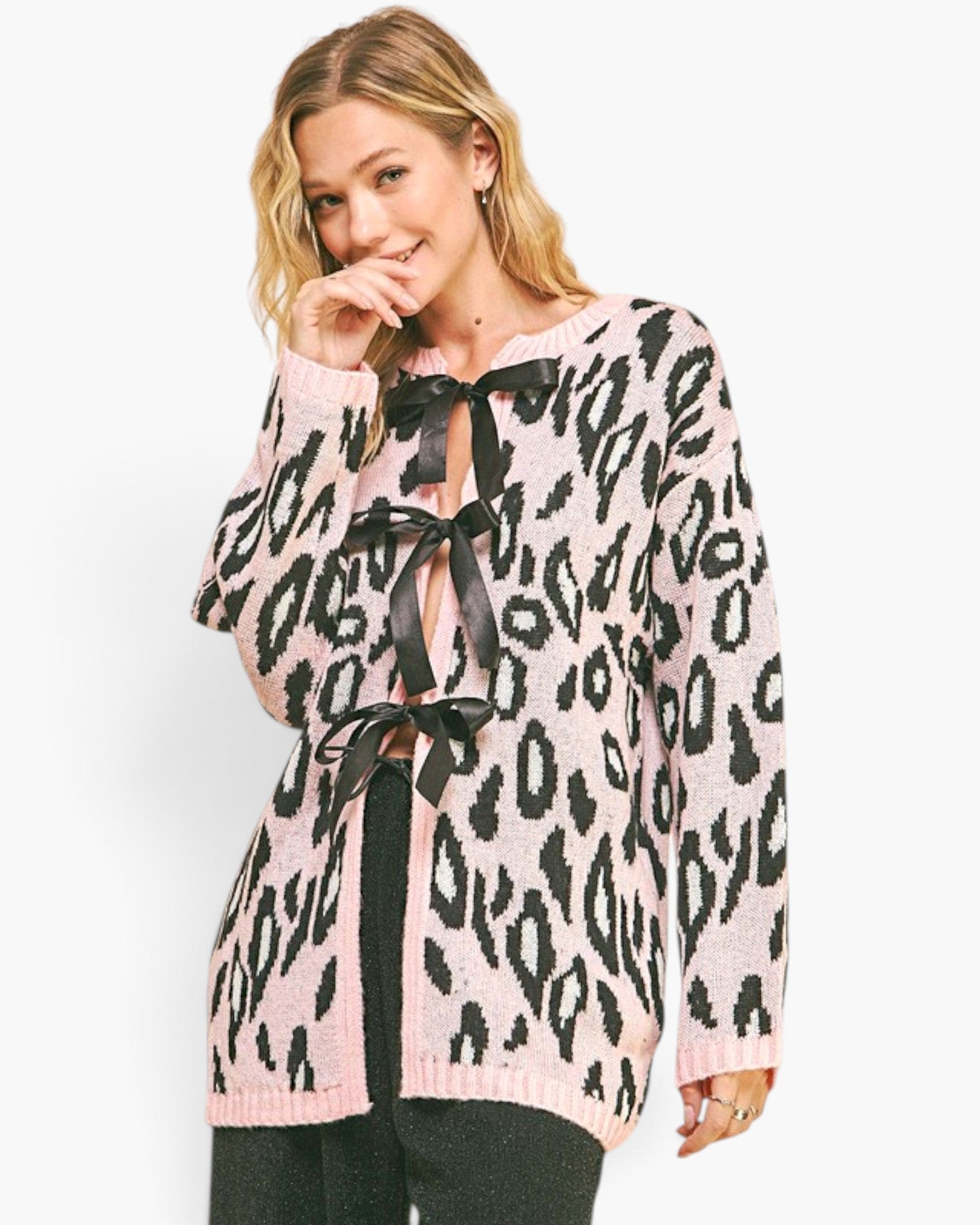 A woman wearing a pink and black leopard print knit cardigan with black ribbon laces, paired with black pants, against a plain white studio background.