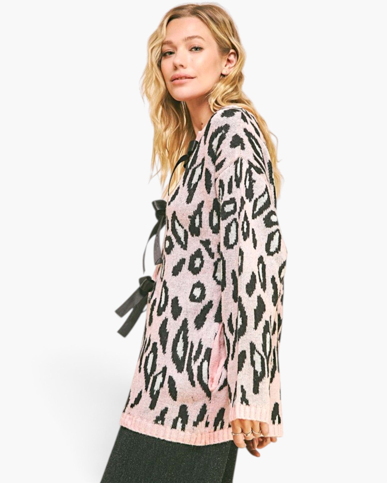A woman wearing a pink and black leopard print knit cardigan with a tie detail at the chest, paired with dark green pants, against a plain white background.
