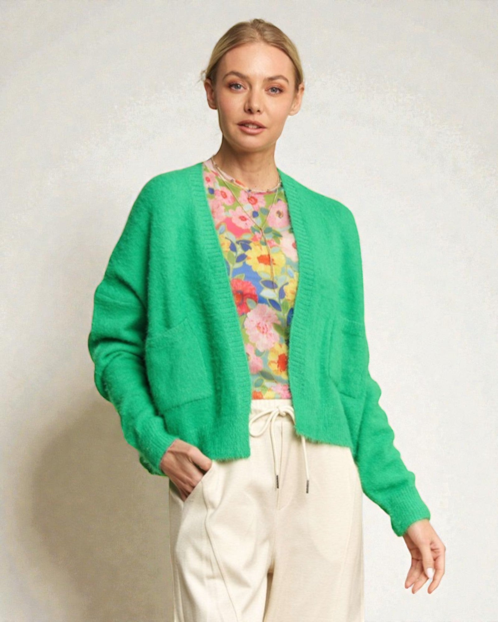 Woman wearing a green cardigan over a floral shirt with white pants on a plain background