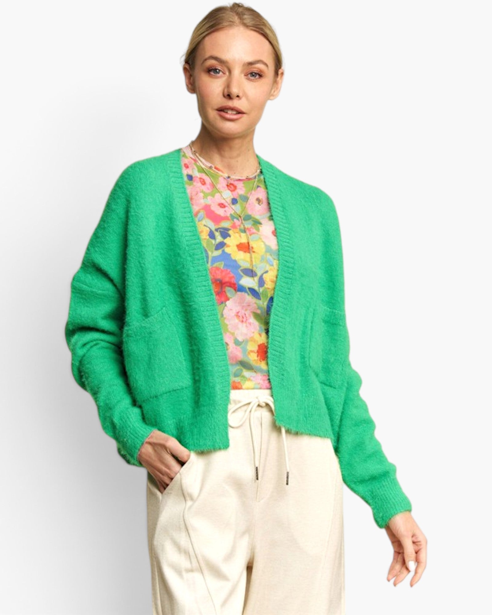 A woman modeling a bright green fuzzy open-front cardigan over a colorful floral top and cream drawstring pants, photographed against a clean light gray studio background.