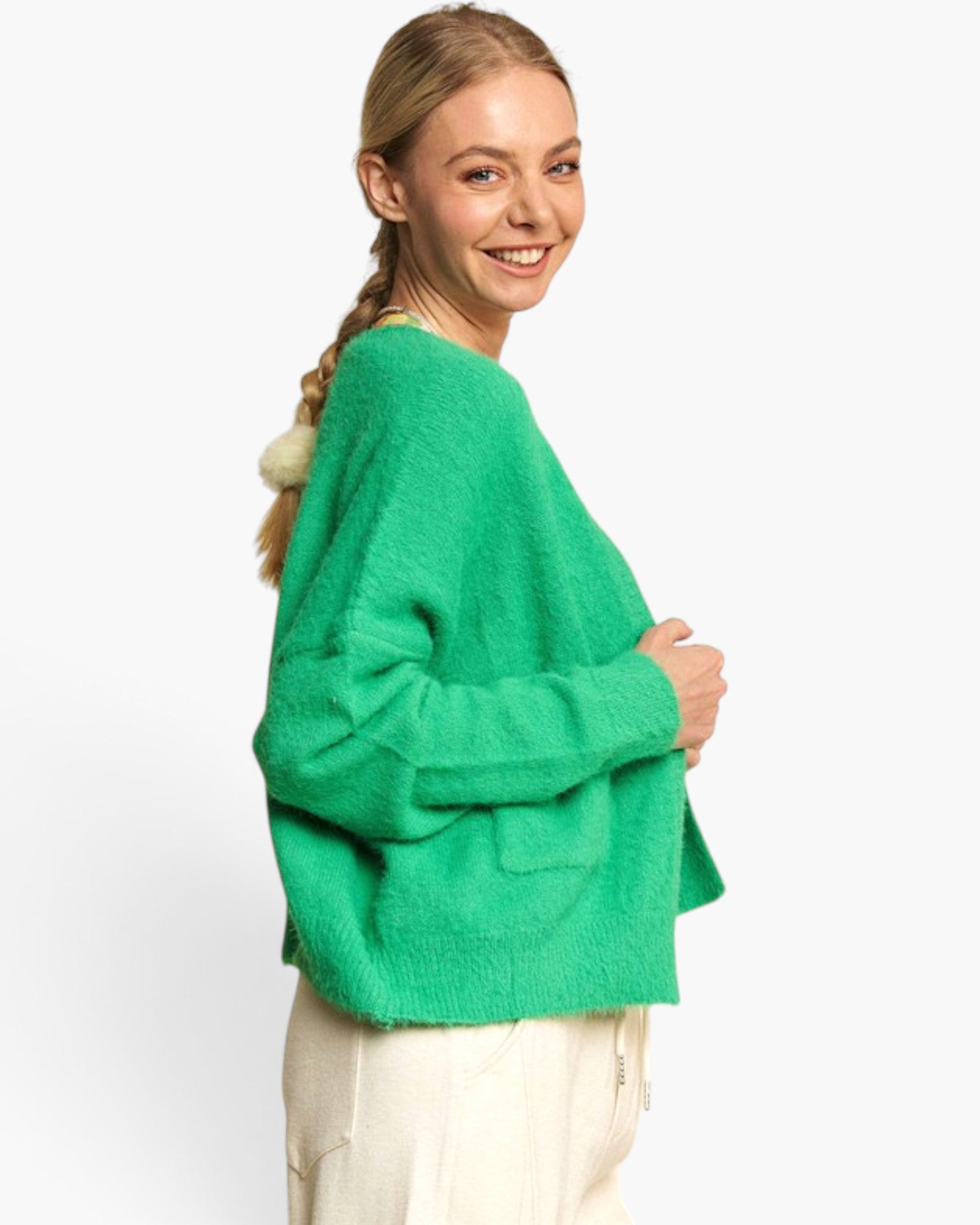 A woman wearing a fuzzy bright green cropped sweater with a pom-pom detail on the back, paired with cream pants, photographed against a clean white studio background.