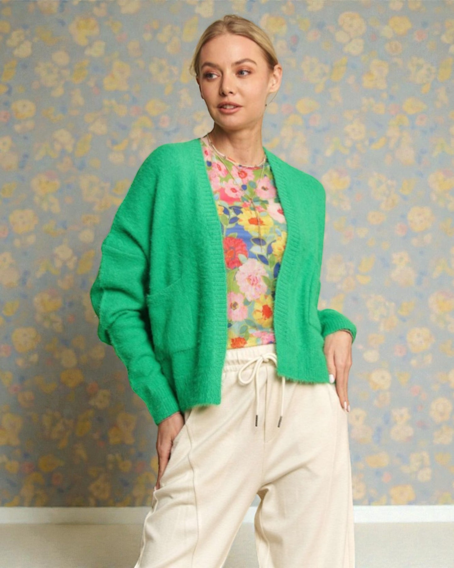 Woman wearing a green cardigan over a floral shirt with a floral-patterned wall background
