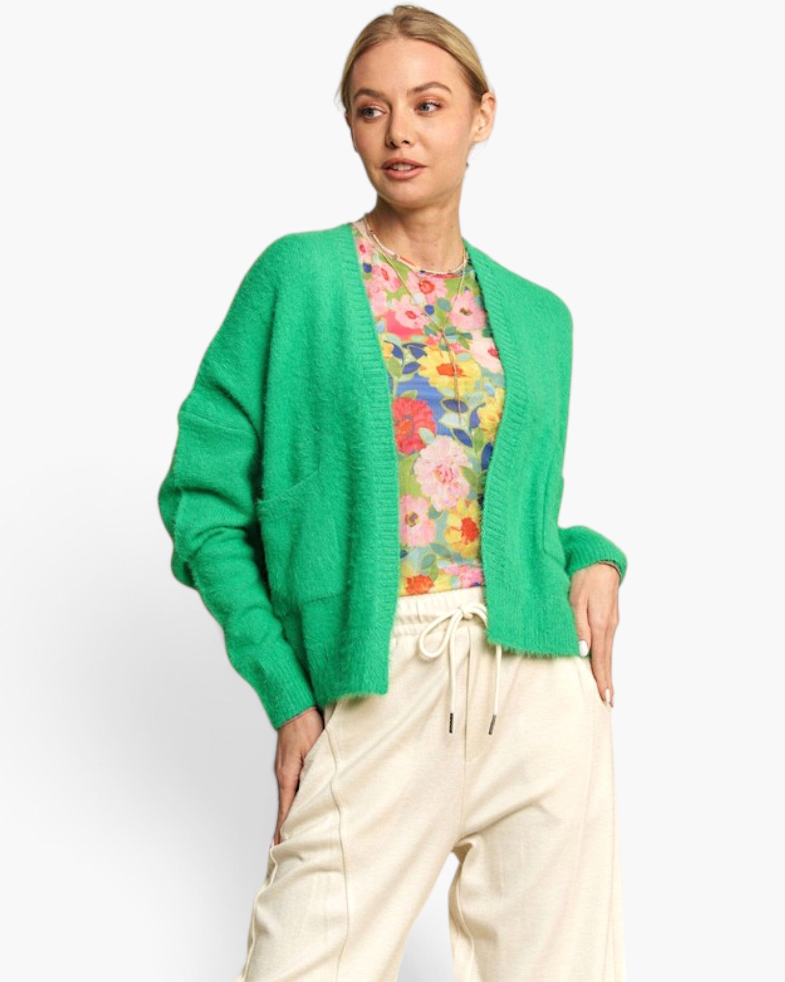 A woman modeling a bright green knit cardigan over a colorful floral top and cream drawstring pants, photographed against a clean light gray studio background.