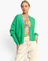 A woman modeling a bright green knit cardigan over a colorful floral top and cream drawstring pants, photographed against a clean light gray studio background.