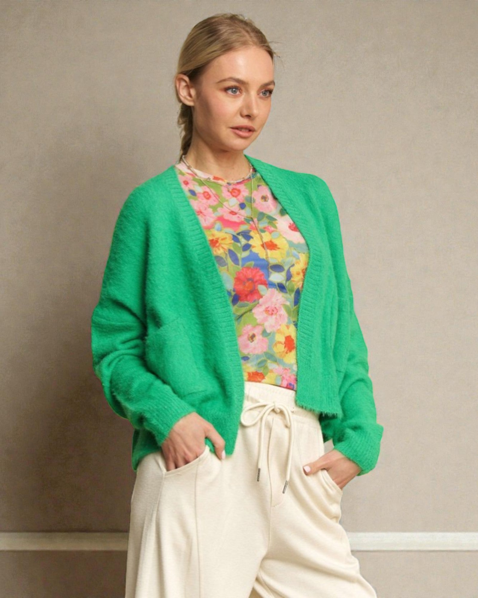 Woman wearing a green cardigan over a floral shirt with white pants against a neutral background