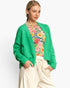 A woman modeling a bright green open-front knit cardigan over a colorful floral top and cream drawstring pants, photographed against a clean light gray studio background.