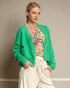 Woman wearing a green cardigan over a floral shirt with white pants against a neutral background