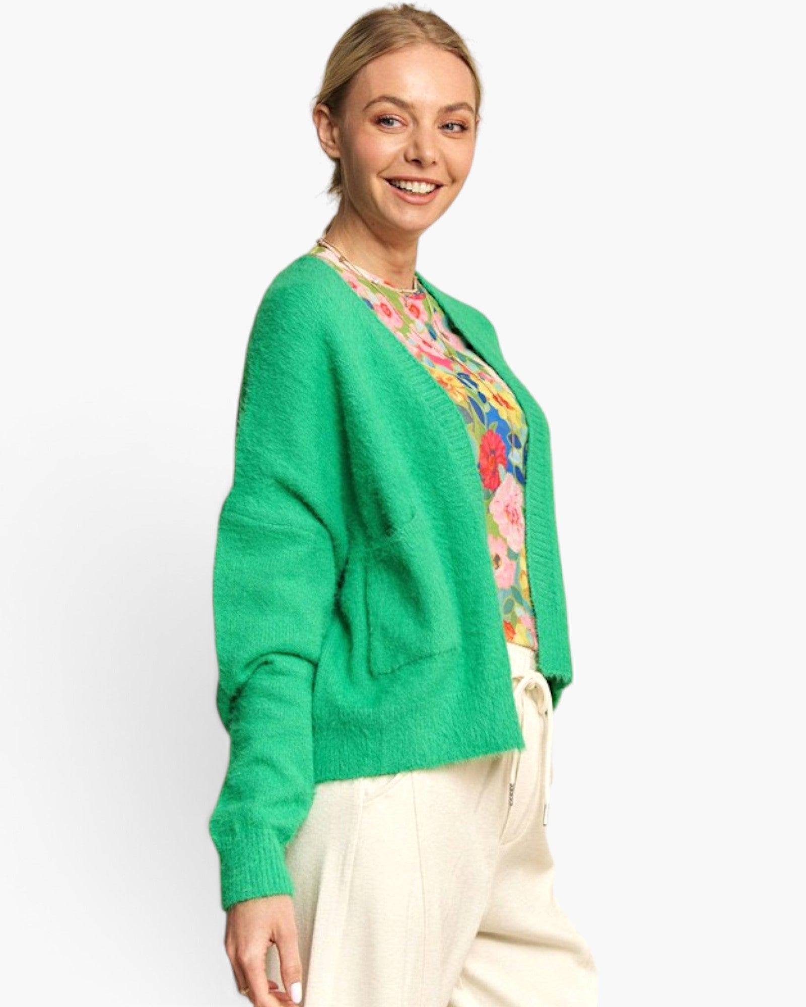 A woman wearing a bright green fuzzy knit cardigan over a colorful floral top and cream pants, photographed against a clean light gray studio background.