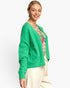 A woman wearing a bright green fuzzy knit cardigan over a colorful floral top and cream pants, photographed against a clean light gray studio background.