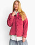 A blonde model wearing a pink quilted corduroy jacket over a white collared shirt and gray skirt, photographed against a plain white studio background.