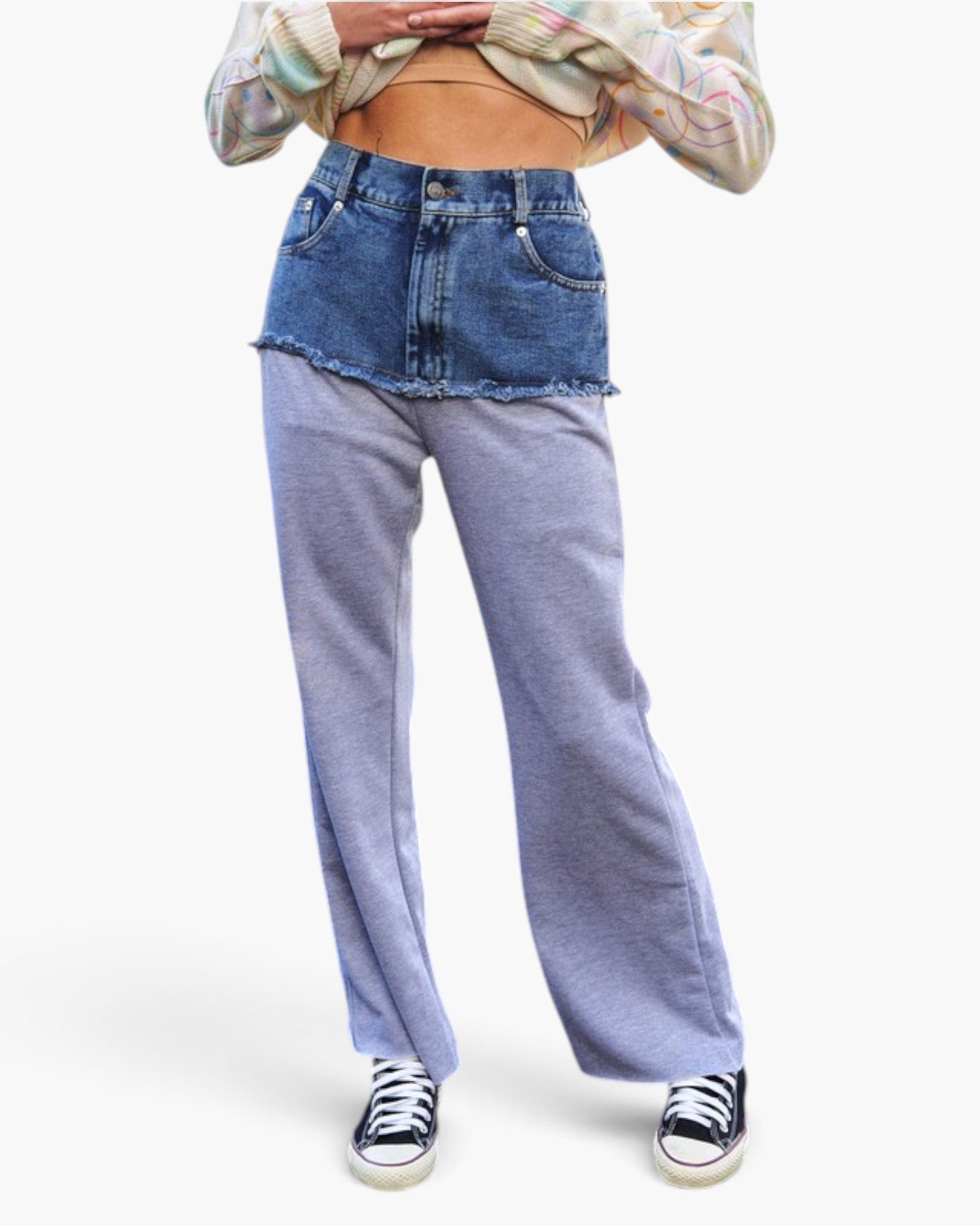 A model is shown from the waist down wearing a pair of wide-leg, color-blocked sweatpants with a blue denim top section and light purple-gray bottom sections, styled with a cropped multicolor shirt and black canvas sneakers.