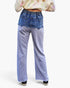 A person shown from the back wearing wide-leg patchwork pants with a heathered light purple lower body and a blue denim back panel, paired with a patterned long-sleeve top and black sneakers against a plain white background.