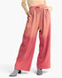 A model wearing wide-leg pink pants with a gradient fade from a deeper rose at the top to a lighter coral-pink at the bottom, decorated with small silver rhinestone-like speckles. The model is cropped from the waist down, wearing a beige top and gray heeled boots, against a plain white studio background.