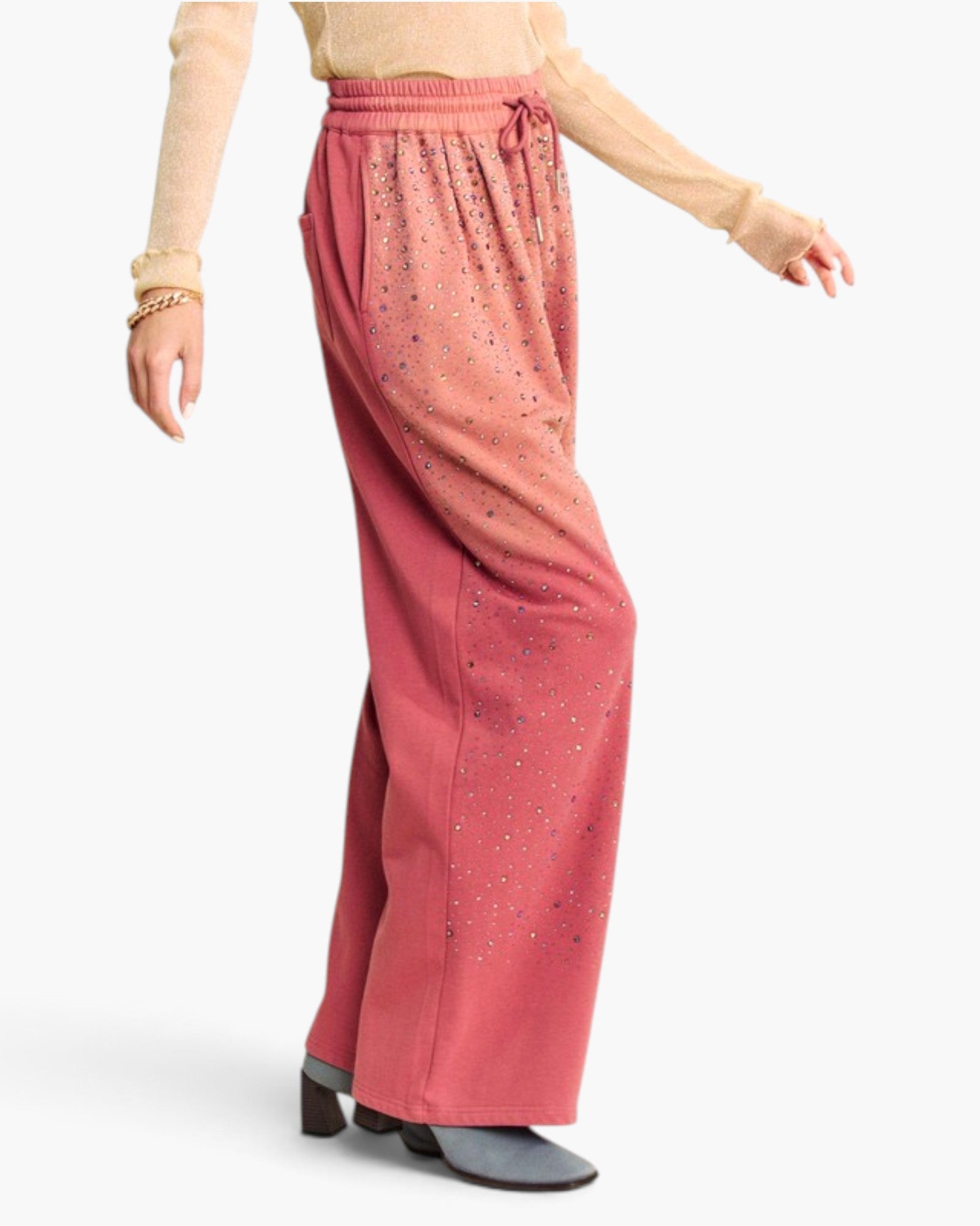 A model wearing wide-leg dusty rose pants with a gradient effect fading to pink at the top and small scattered rhinestone embellishments, paired with a beige long-sleeve top and gray heeled boots against a white studio background.