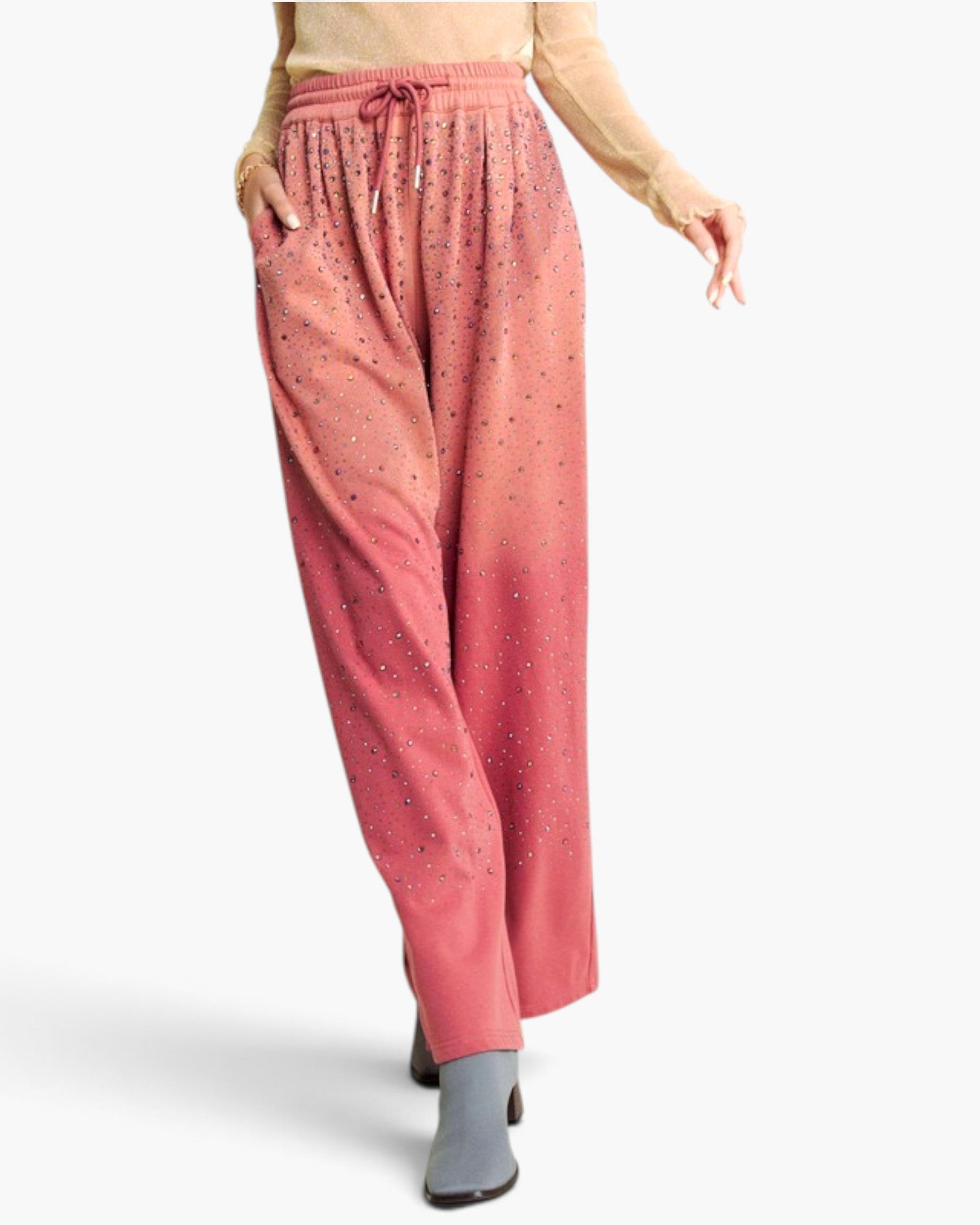 A model is shown from the waist down wearing wide-leg, relaxed-fit pants with a pink and coral ombre gradient and small rhinestone embellishments. The pants feature an elastic drawstring waistband and side pockets, paired with a beige long-sleeve top and gray ankle boots against a plain white studio background.