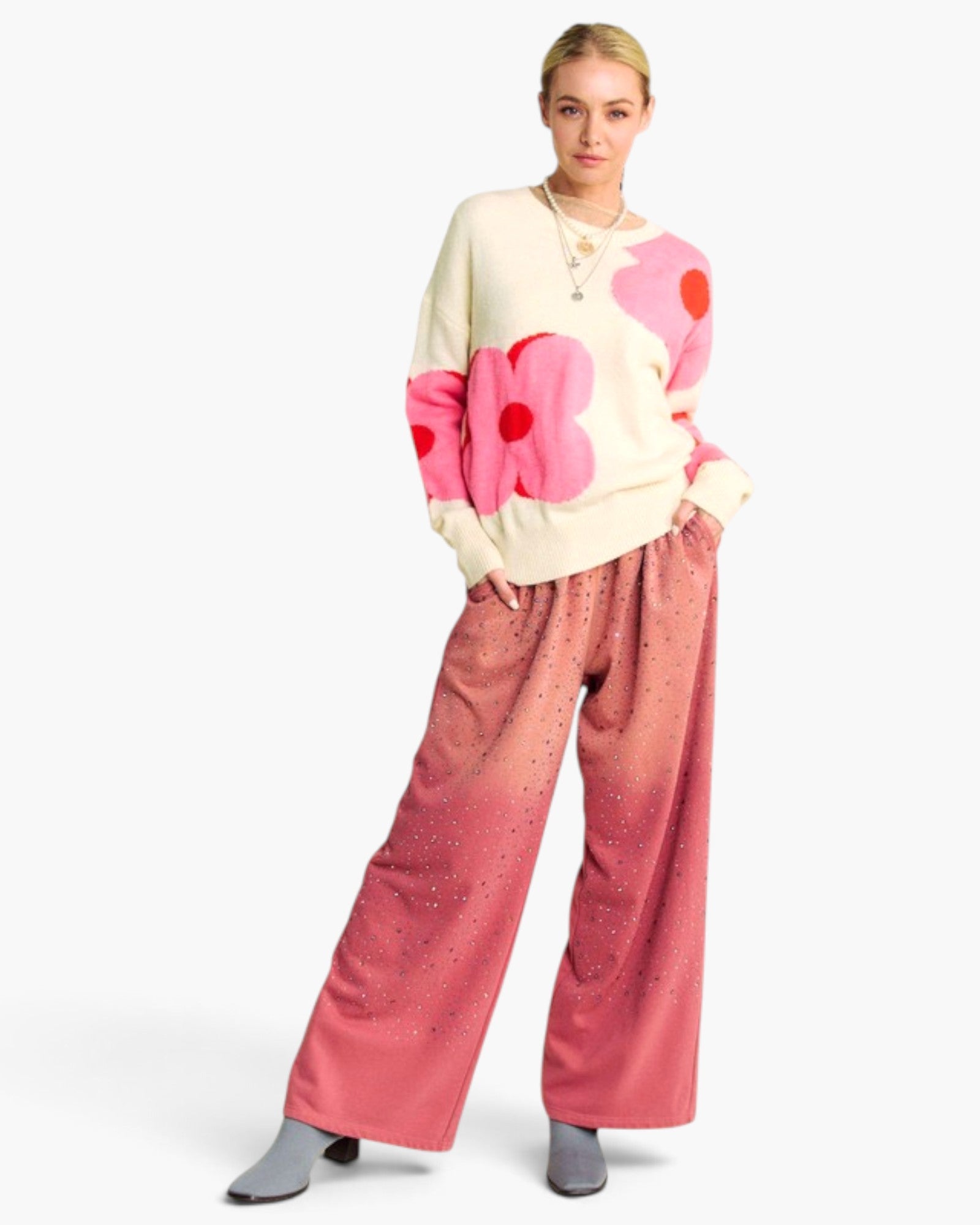 A model wearing a cream knit sweater with large pink and red flower motifs, paired with wide-leg pink gradient pants featuring small silver rhinestone embellishments, and gray pointed-toe boots, against a plain white background.