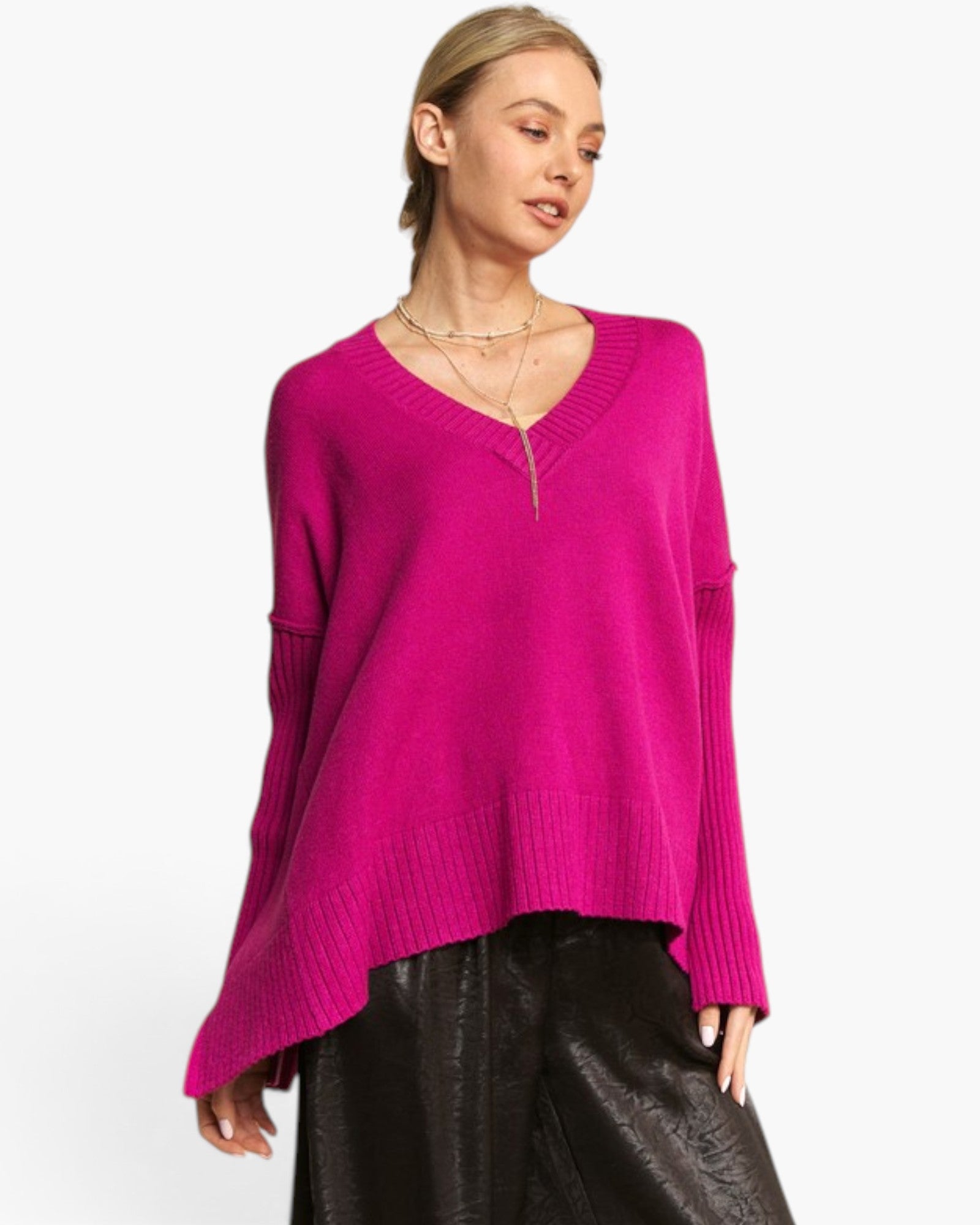 A woman wearing a bright magenta pink knit sweater with a V-neck and drop shoulders, paired with black leather pants, against a plain white background.