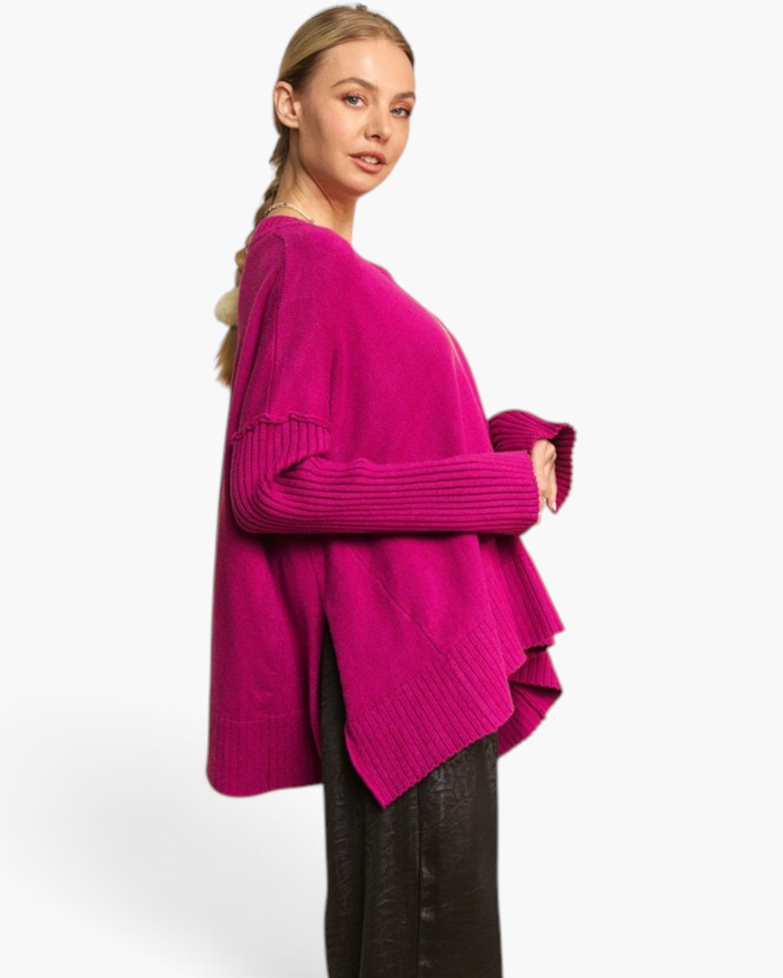 A woman modeling a bright magenta knit sweater with a relaxed fit, ribbed cuffs, and an asymmetrical layered hem, paired with dark pants against a plain light gray studio background.