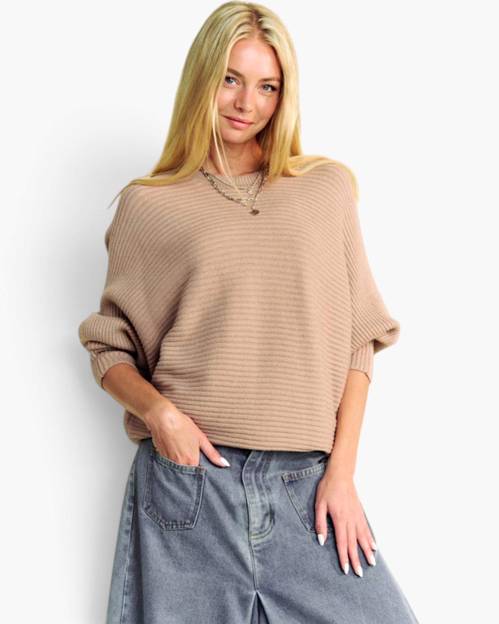 A blonde woman wearing a beige ribbed knit sweater with elbow-length sleeves and a relaxed fit, paired with light-wash denim jeans, against a plain white studio background.