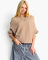 A blonde woman wearing a beige ribbed knit sweater with elbow-length sleeves and a relaxed fit, paired with light-wash denim jeans, against a plain white studio background.