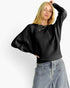 A woman wearing a black ribbed knit long-sleeve sweater with wide batwing sleeves and light-wash blue jeans, posed against a plain white studio background.
