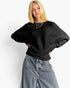 A blonde woman wearing a black ribbed knit top with wide batwing sleeves and light-wash wide-leg jeans, standing against a plain white studio background.