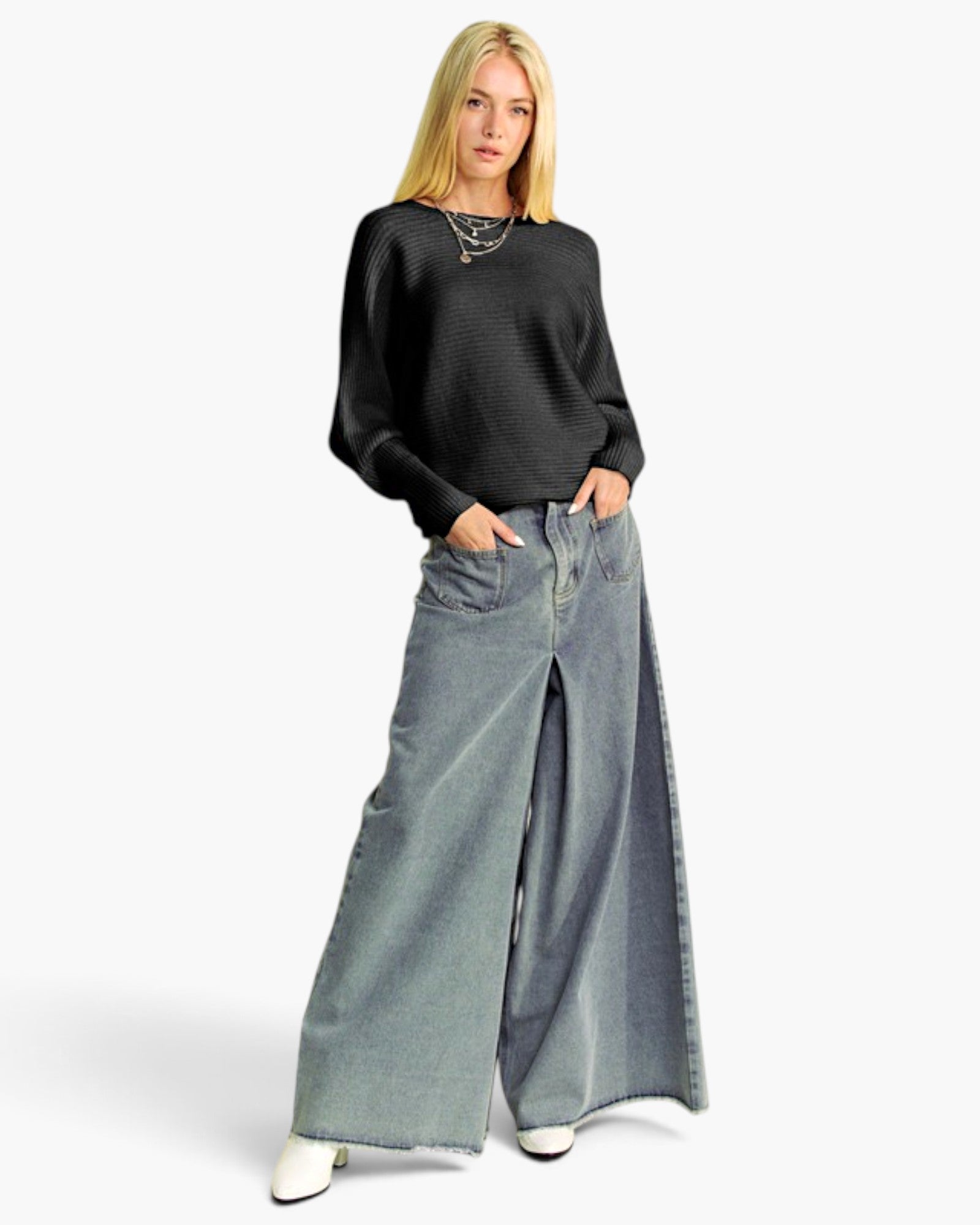 A blonde model wearing a black ribbed knit long-sleeve top and wide-leg light blue denim pants with side slits, standing against a light gray studio background.