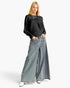 A blonde model wearing a black ribbed knit long-sleeve top and wide-leg light blue denim pants with side slits, standing against a light gray studio background.