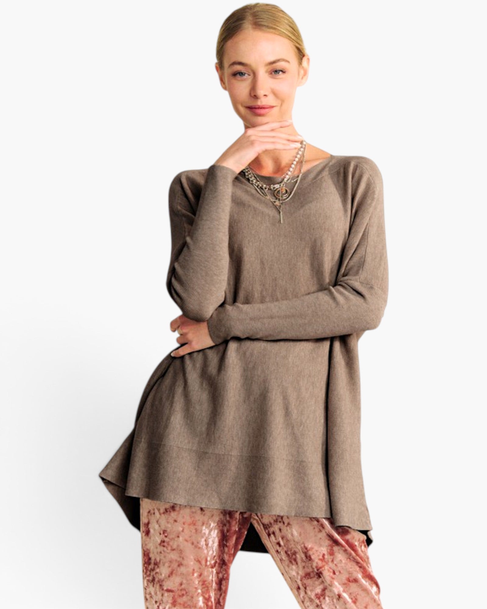 A woman modeling a taupe brown long-sleeve knit tunic top with a high-low hem, paired with patterned pink pants, against a plain white studio background.