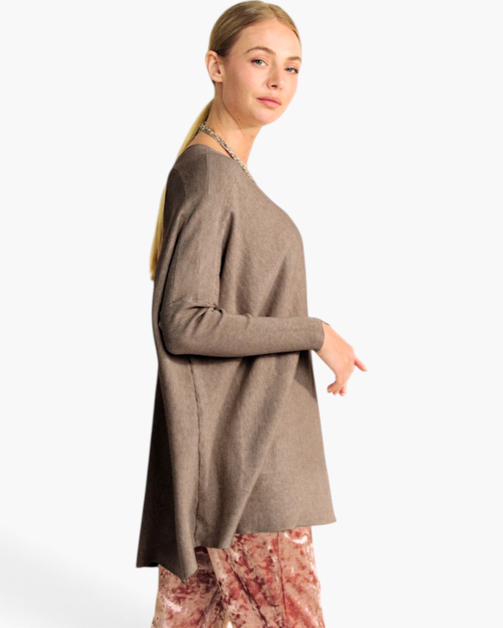 A woman modeling a taupe brown long-sleeve knit top with a wide, draped silhouette, paired with patterned pink pants, against a plain white studio background.