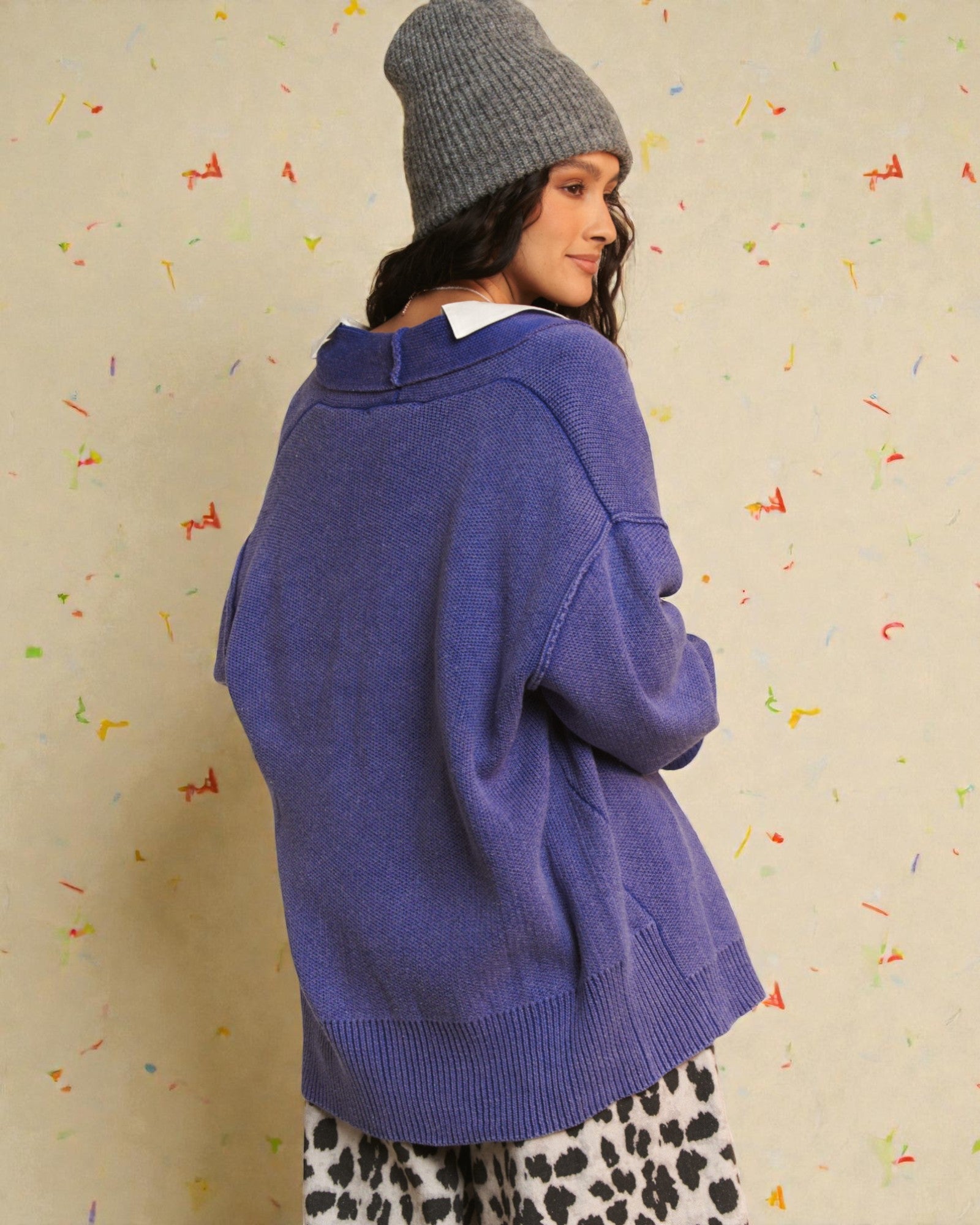 Person wearing a blue sweater and gray beanie against a colorful confetti-patterned wall.