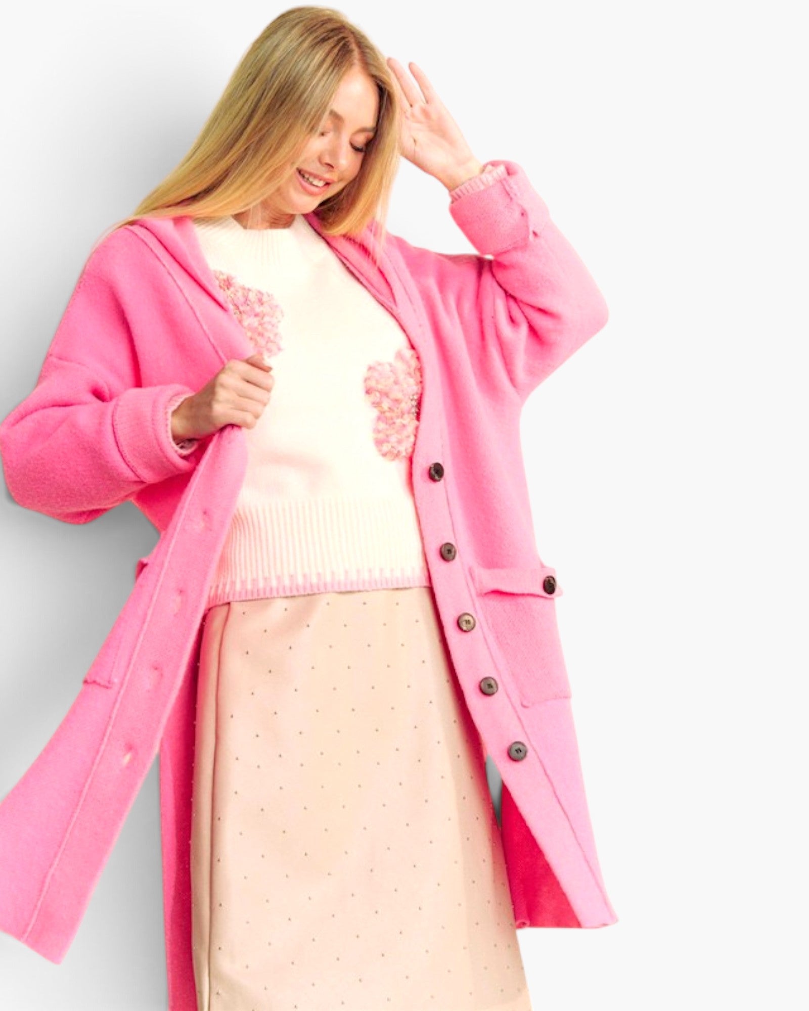 A woman wearing a bright pink open-front cardigan over a cream sweater and a light pink skirt, posed against a plain light gray studio background.