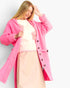 A woman wearing a bright pink open-front cardigan over a cream sweater and a light pink skirt, posed against a plain light gray studio background.