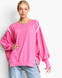 A woman modeling a bright pink oversized crewneck sweatshirt with balloon sleeves and light pink piping details along the shoulders and sides, paired with light blue jeans against a plain white background.