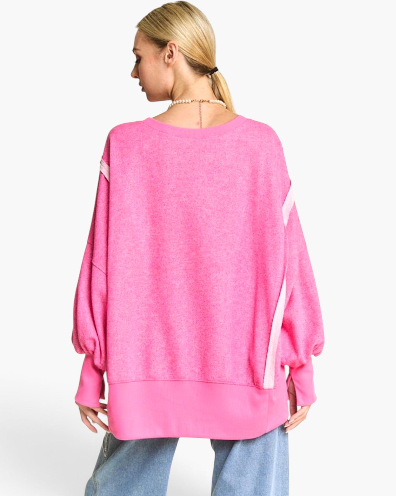 A woman shown from the back wearing a bright pink oversized sweatshirt with white diagonal stripe accents on the sleeves, paired with light blue jeans, against a plain white studio background.