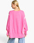 A woman shown from the back wearing a bright pink oversized sweatshirt with white diagonal stripe accents on the sleeves, paired with light blue jeans, against a plain white studio background.