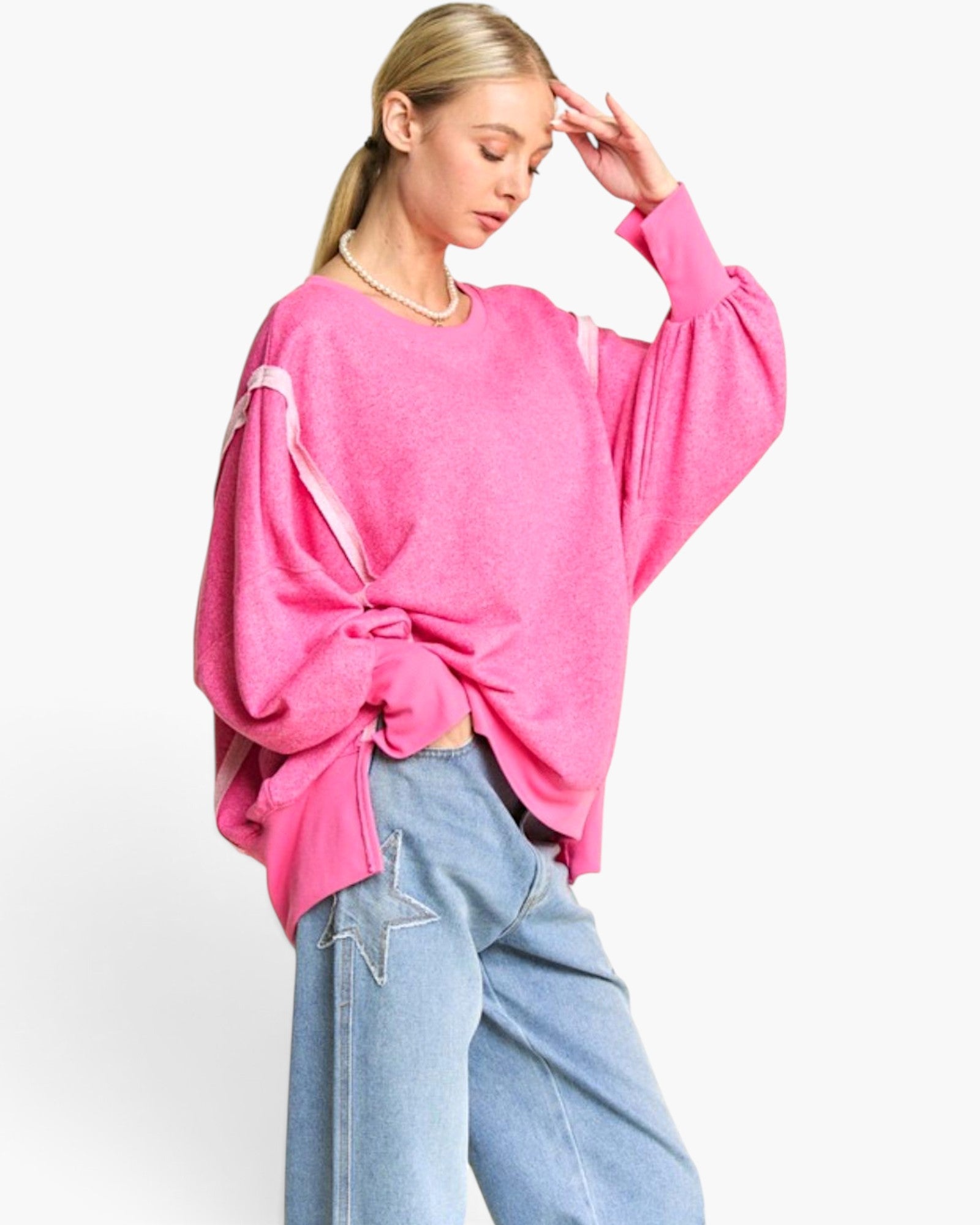 A woman modeling a bright pink oversized knit sweater with balloon sleeves and a twisted knot detail at the waist, paired with light blue jeans against a plain white background.