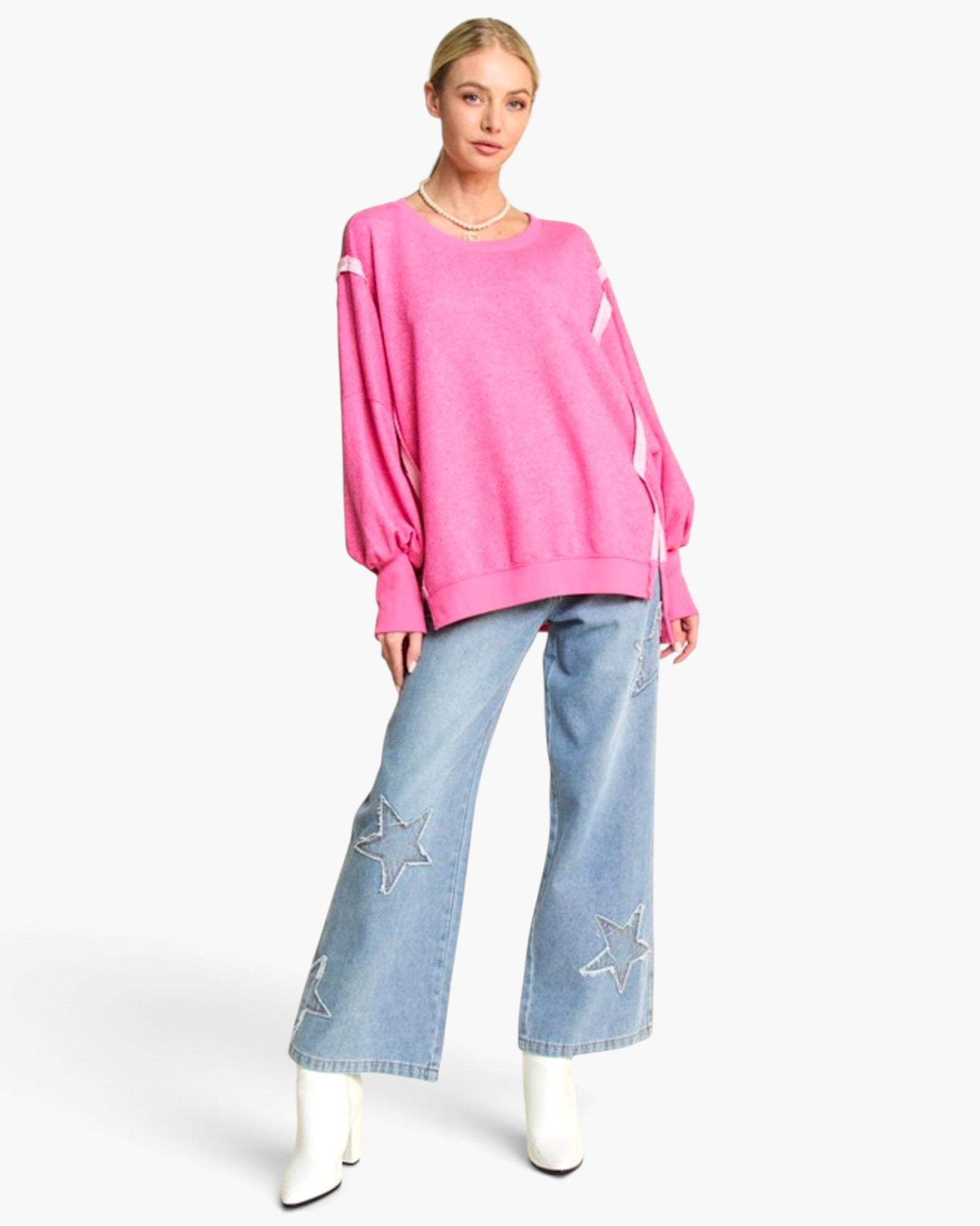 A model wearing a bright pink crewneck sweater with white stripe accents on the sleeves, paired with light blue wide-leg jeans featuring star embroidery and white pointed boots, set against a plain white studio background.