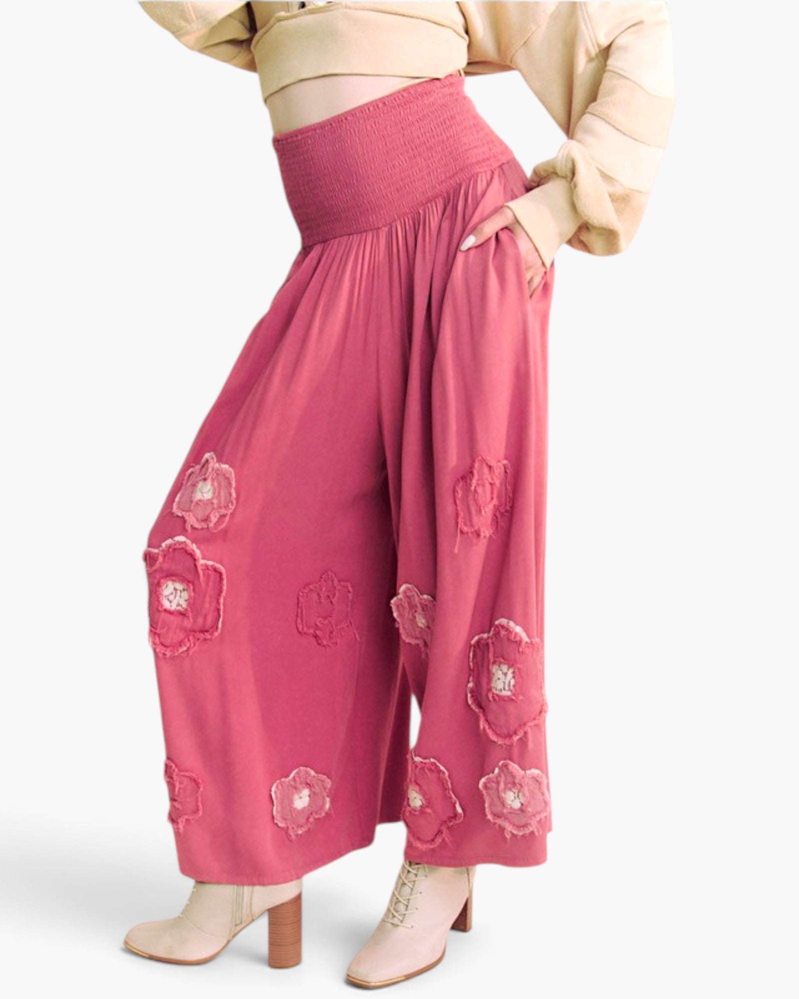 A model wearing high-waisted, wide-leg pink pants with 3D floral embroidery and sequin details, paired with a beige cropped top and beige heels against a light gray background.