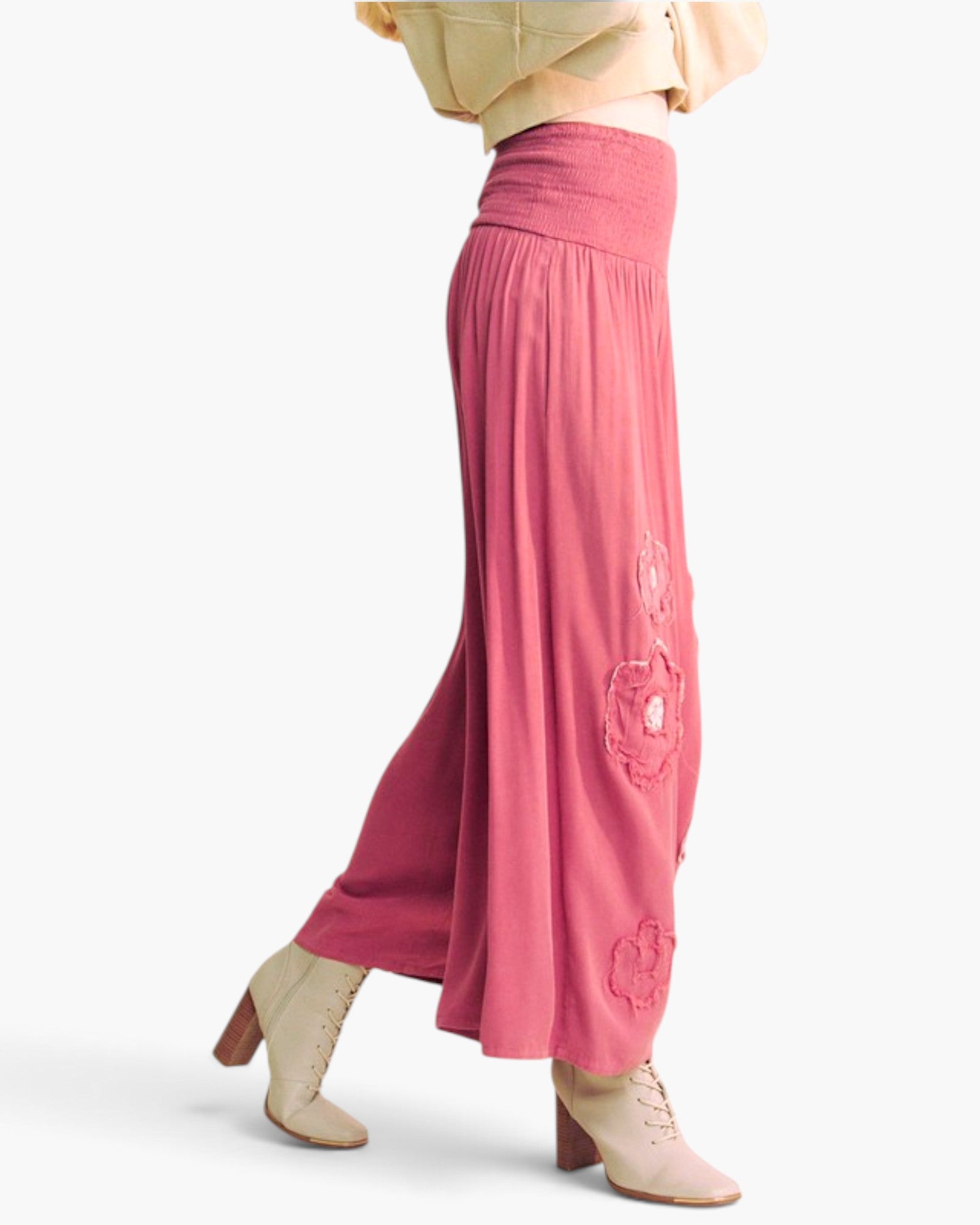 A model is shown from the waist down wearing a pair of high-waisted, wide-leg pink pants with tonal floral embroidery down the side, paired with beige ankle boots against a light gray studio background.